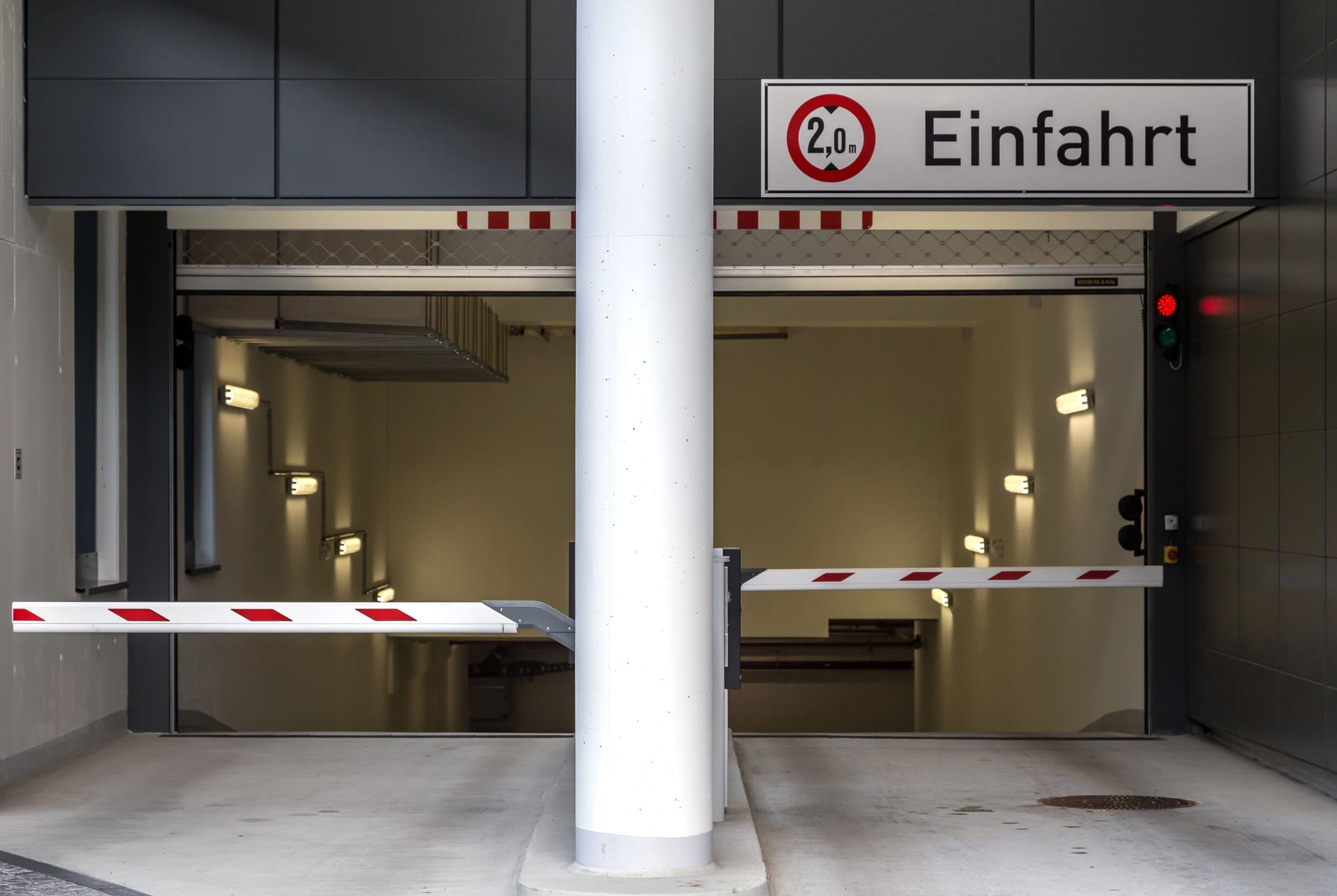 Die Einfahrt zu einer Tiefgarage, gekennzeichnet durch eine rote Ampel, eine weiße Säule und eine geschlossene weiße Schranke mit roten Streifen. Über dem Eingang hängt ein Schild mit der Aufschrift '2,0 m Einfahrt'.