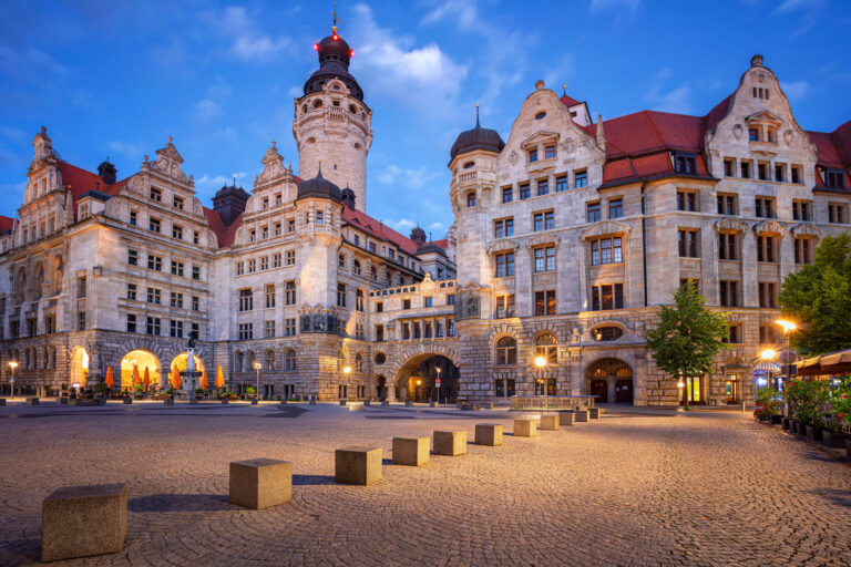 Abendaufnahme des Neuen Rathauses in Leipzig: Ein historisches Steingebäude im Renaissance-Stil mit einem hohen Turm, dessen Spitze rot leuchtet. Der gepflasterte Rathausplatz im Vordergrund wird durch Bodenlampen und warme Fensterbeleuchtung erhellt.