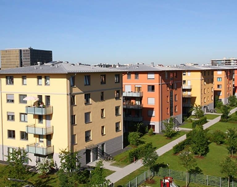 Mehrere vierst&ouml;ckige, modern aussehende Mehrfamilienh&auml;user in den Farben Gelb, Orange und Dunkelrot, umgeben von gepflegten Rasenfl&auml;chen und Fu&szlig;wegen unter blauem Himmel.
