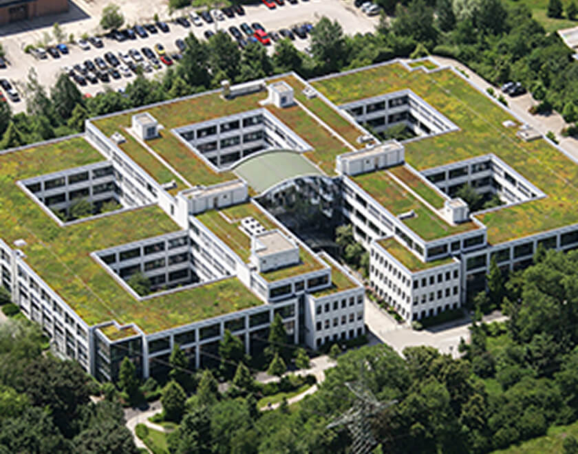 Luftaufnahme eines großen, modernen Bürokomplexes mit weißen Fassaden, das eine komplexe, rechteckige Struktur mit mehreren Innenhöfen bildet. Das Flachdach ist vollständig begrünt (Gründach). Im Hintergrund ist ein großer Parkplatz sichtbar.