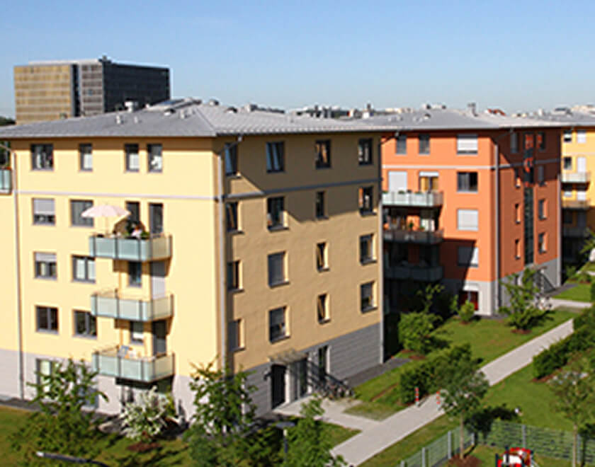 Mehrere vierstöckige, modern aussehende Mehrfamilienhäuser in den Farben Gelb, Orange und Dunkelrot, umgeben von gepflegten Rasenflächen und Fußwegen unter blauem Himmel.