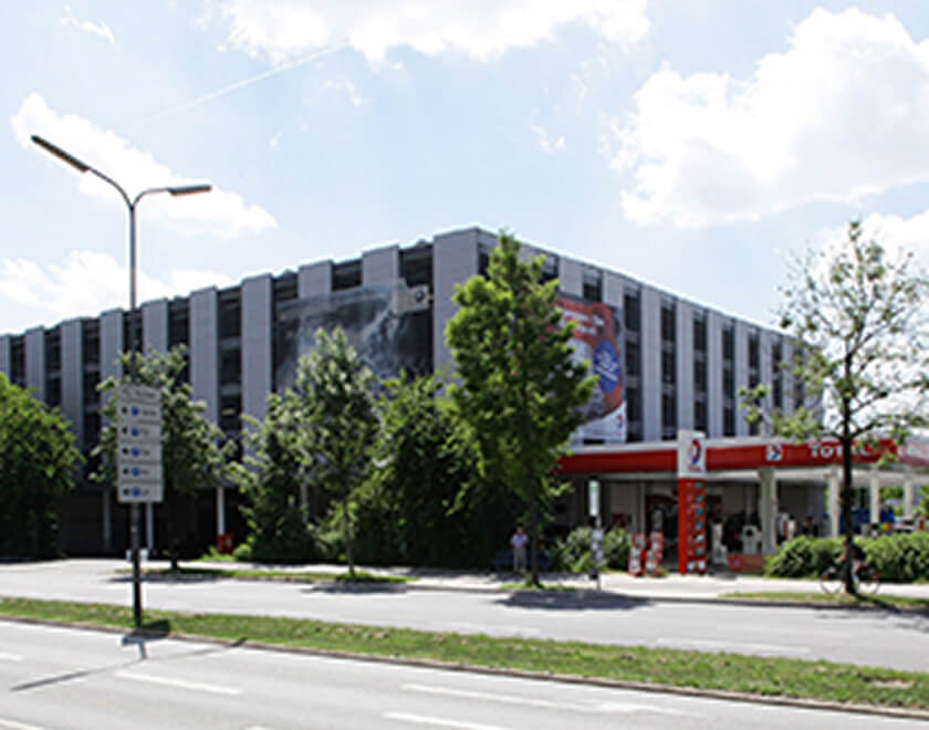 Dunkles, mehrstöckiges Parkhaus mit vertikalen Streifen, das großflächige Werbebanner an der Fassade trägt. Im Vordergrund, rechts neben dem Parkhaus, befindet sich eine moderne, rot-weiß gestaltete Tankstelle.