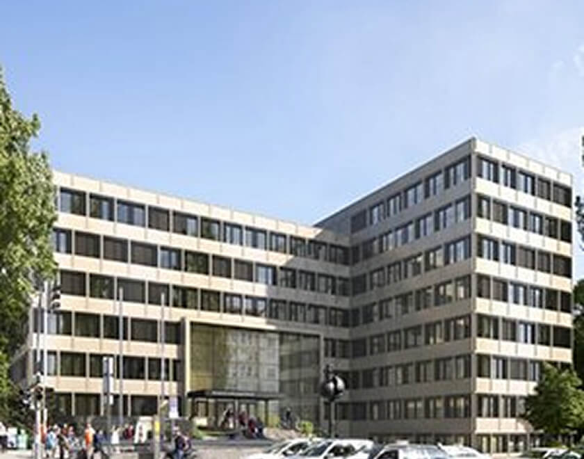 Großes, sechsgeschossiges Büro- oder Verwaltungsgebäude im Stil der 1960er Jahre mit einer beigefarbenen Steinfassade und horizontalen Fensterreihen. Der Haupteingang in der Mitte ist zurückgesetzt und verglast. Vor dem Gebäude sind Fußgänger und parkende Autos auf der Straße zu sehen.