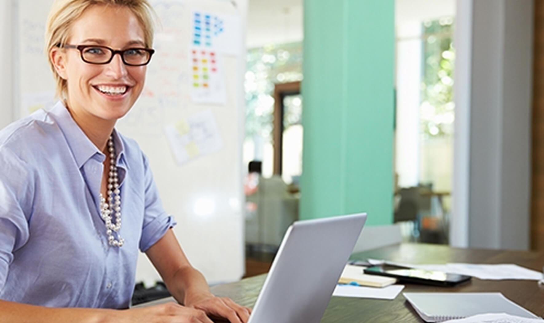 Eine lächelnde Frau mit Brille und kurzer blonder Frisur arbeitet an einem silbernen Laptop in einem hellen, modernen Büro. Sie trägt ein hellblaues Hemd und eine lange Perlenkette.