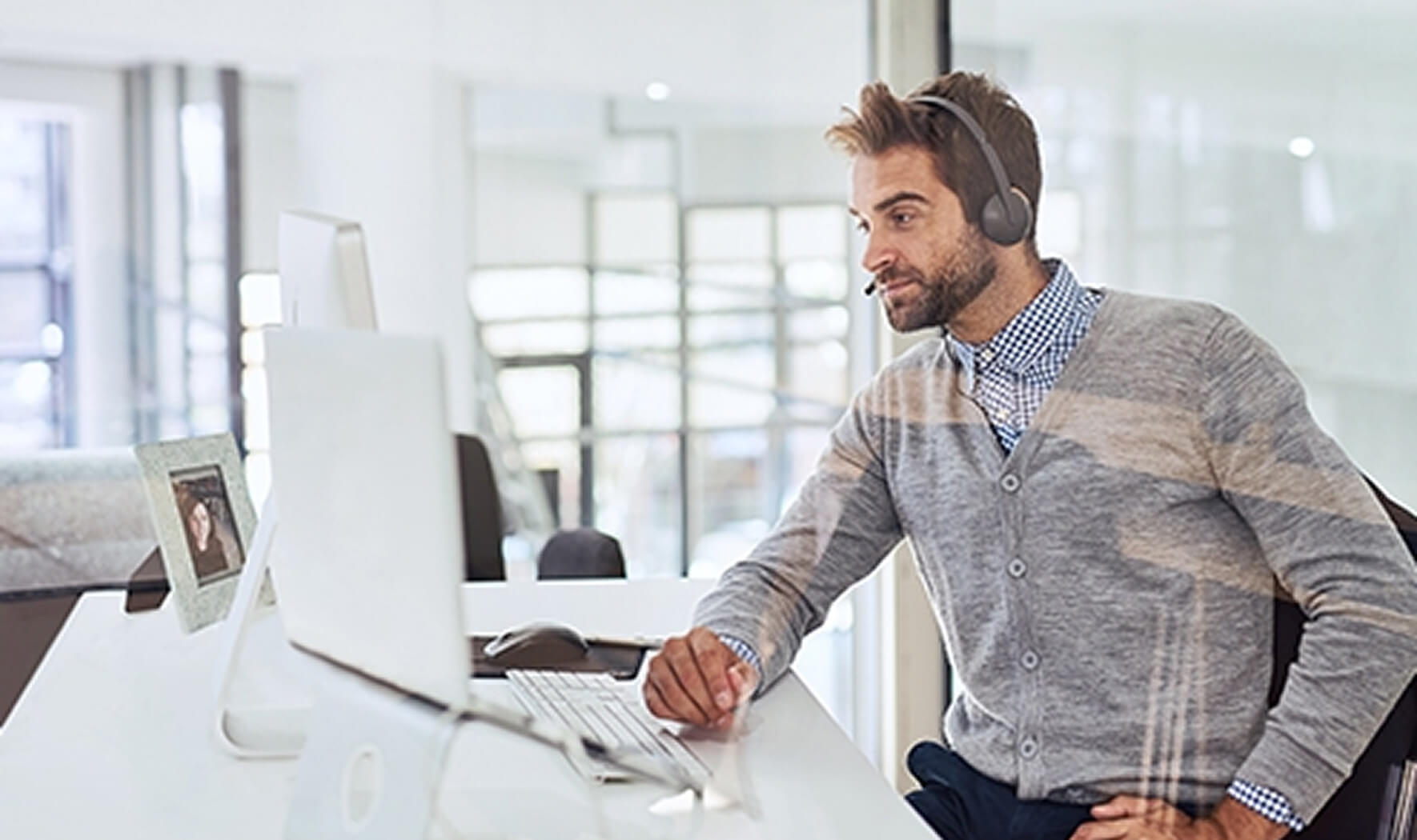 Ein lächelnder Mann in einem grauen Cardigan sitzt an einem Schreibtisch in einem hellen Büro und arbeitet an einem Computer. Er trägt ein Headset mit Mikrofon und sieht konzentriert auf den Bildschirm.