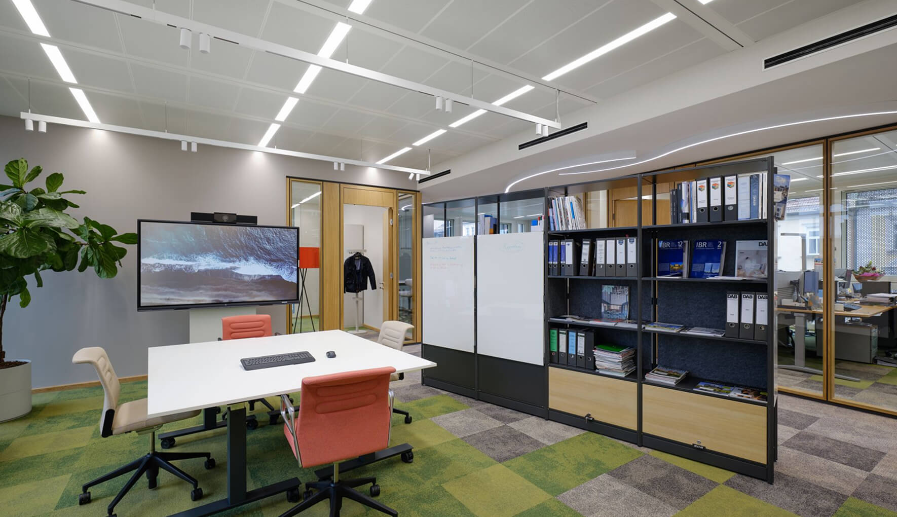 Moderner Besprechungsraum in einem Büro mit einem rechteckigen weißen Tisch und vier Bürostühlen. Eine große Videowall mit einem Bild von Wellen hängt an der Wand. Der Raum ist durch ein hohes Regal mit Aktenordnern und zwei Whiteboards vom Großraumbüro getrennt.