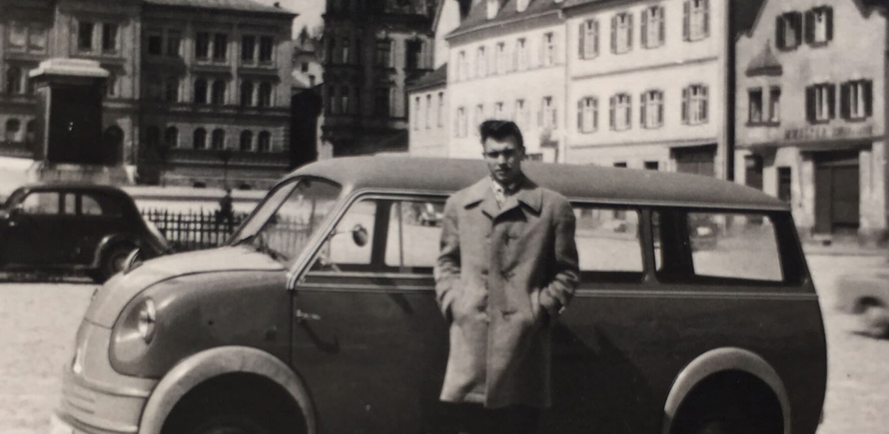 Schwarz-Weiß-Foto: Ein junger Mann in einem Trenchcoat steht lässig vor einem kleinen, rundlichen Kleinstwagen der Nachkriegszeit, der wahrscheinlich ein Messerschmitt oder ein ähnliches Modell ist. Im Hintergrund sind historische, mehrstöckige Gebäude an einem Platz zu sehen.