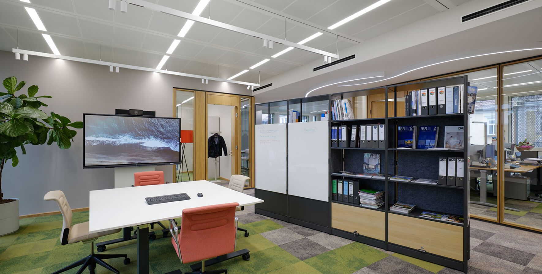 Moderner Besprechungsraum in einem Büro mit einem rechteckigen weißen Tisch und vier Bürostühlen. Eine große Videowall mit einem Bild von Wellen hängt an der Wand. Der Raum ist durch ein hohes Regal mit Aktenordnern und zwei Whiteboards vom Großraumbüro getrennt.
