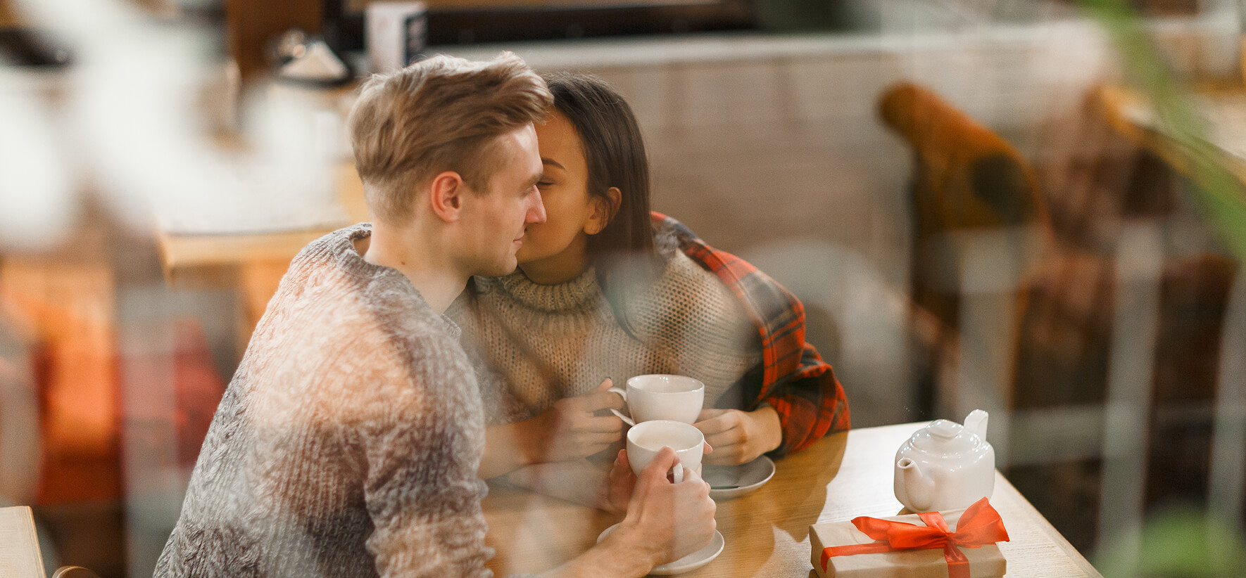 Ein junges Paar sitzt in einem Café an einem Holztisch, trinkt Kaffee oder Tee und tauscht Zärtlichkeiten aus. Ein Mann im grauen Pullover hält eine Tasse, während eine Frau im karierten Hemd ihm zärtlich die Wange küsst. Rechts liegt ein Geschenk mit roter Schleife.