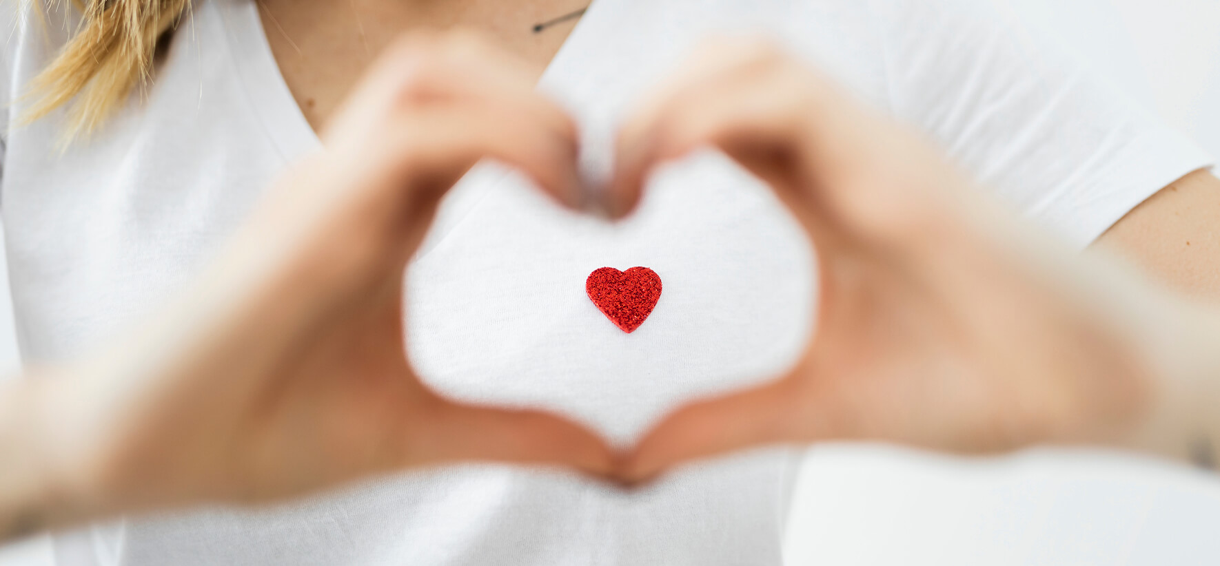 Eine Person in einem weißen T-Shirt formt mit beiden Händen eine Herz-Geste, in deren Mitte ein kleines, glitzerndes, rotes Herz abgebildet ist. Die Hände sind leicht unscharf, um das rote Herz zu fokussieren.