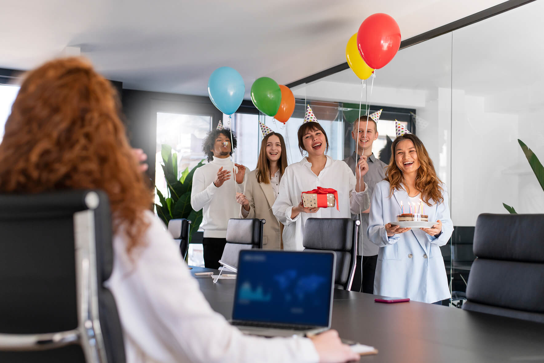 Eine Gruppe von fünf lächelnden Kollegen in Partyhüten überrascht eine Kollegin, deren Rücken zum Betrachter gewandt ist, in einem Büro mit einem Geburtstagskuchen, einem Geschenk und bunten Luftballons. Die überraschte Person sitzt an einem Schreibtisch vor einem Laptop.