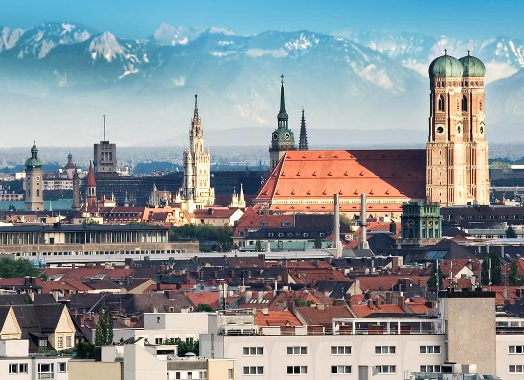 Panoramablick auf die Münchner Altstadt mit einer Dächerlandschaft aus roten Ziegeln im Vordergrund und bekannten Türmen, darunter das Neue Rathaus und die Frauenkirche. Im Hintergrund erheben sich die schneebedeckten Gipfel der Alpen, die durch einen blauen Dunst scheinen.