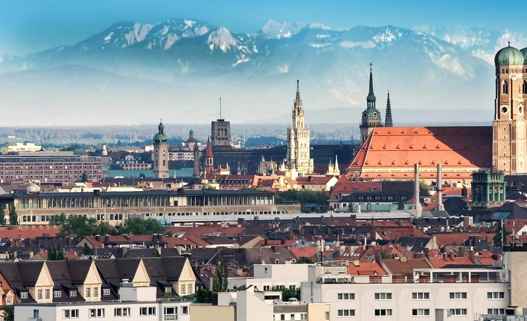 Panoramablick auf die Münchner Altstadt mit einer Dächerlandschaft aus roten Ziegeln im Vordergrund und bekannten Türmen, darunter das Neue Rathaus und die Frauenkirche. Im Hintergrund erheben sich die schneebedeckten Gipfel der Alpen, die durch einen blauen Dunst scheinen.