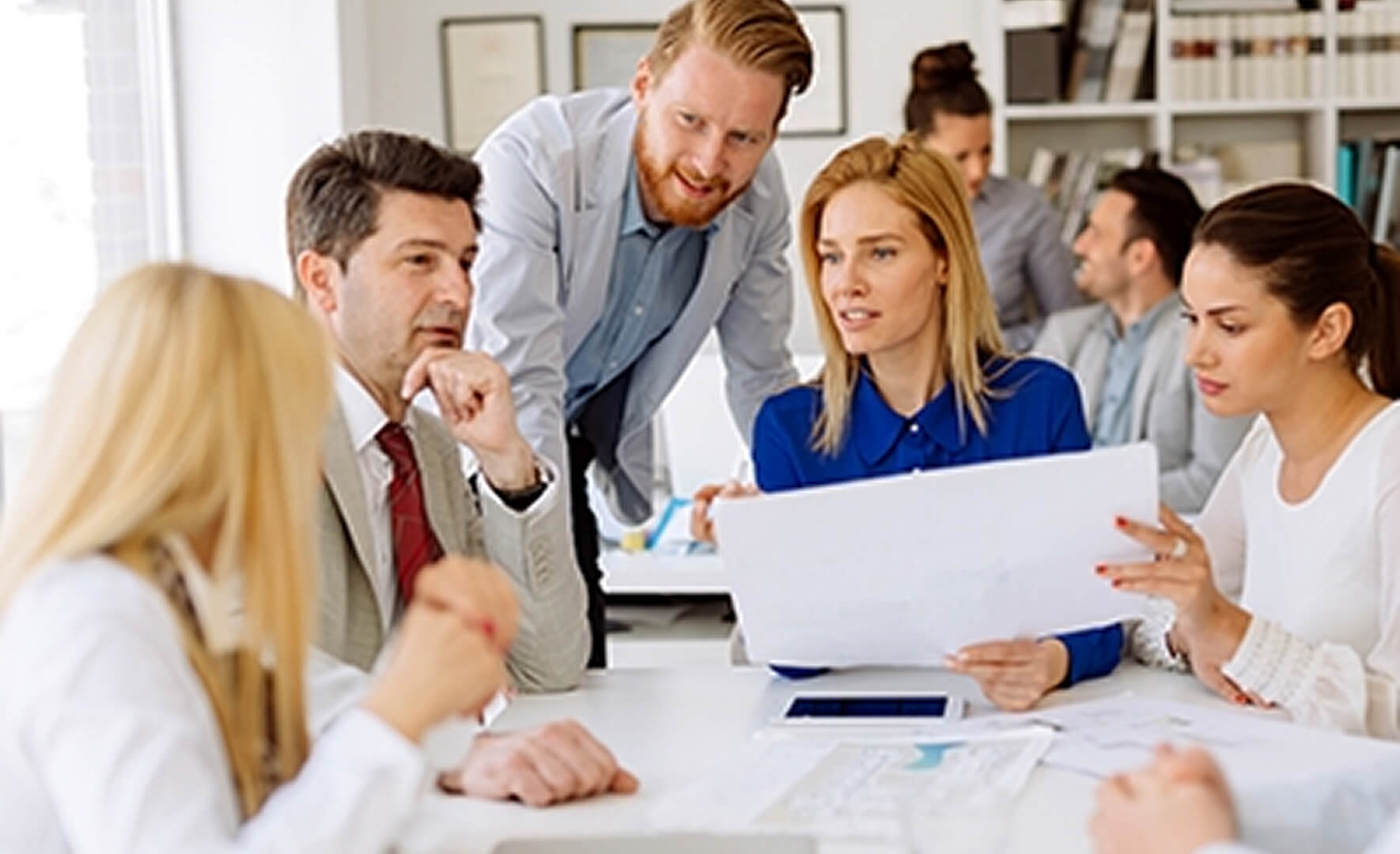 Fünf Geschäftsleute sitzen oder stehen um einen Konferenztisch herum und diskutieren Dokumente während eines Meetings in einem modernen Büro. Eine blonde Frau in einem blauen Hemd hält ein weißes Blatt Papier in der Mitte.