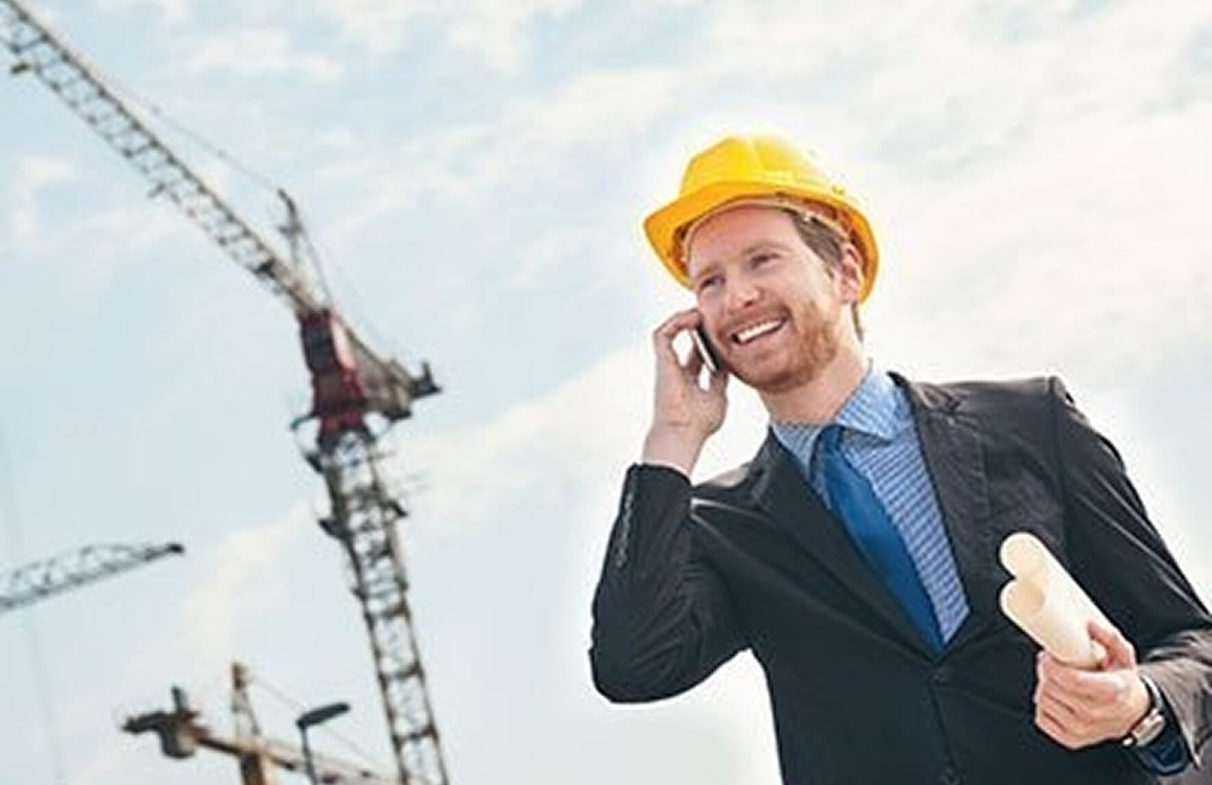 Ein lächelnder Ingenieur oder Bauleiter in einem Anzug mit blauem Schlips und gelbem Schutzhelm telefoniert. Er hält Baupläne in der Hand. Im Hintergrund sind mehrere große Kräne auf einer Baustelle vor einem bewölkten Himmel sichtbar.