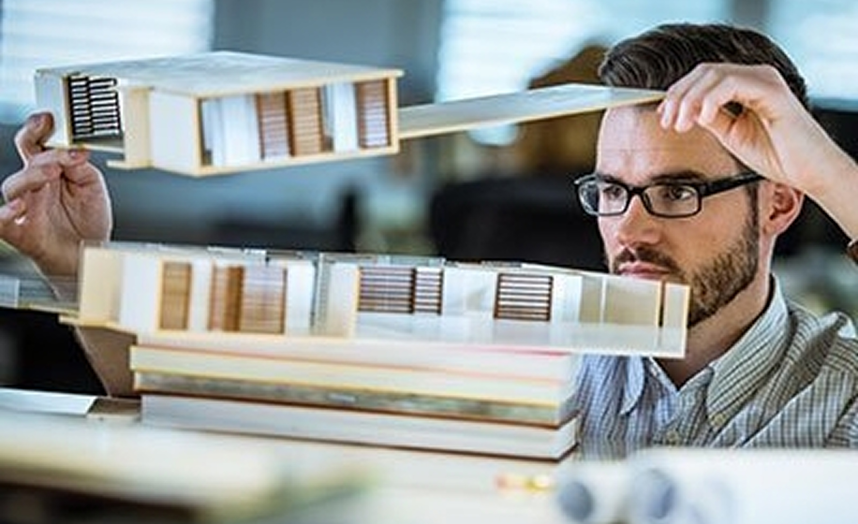 Ein Architekt mit Brille hält und untersucht konzentriert ein detailreiches Holzmodell eines modernen Gebäudes, das er gerade zusammenbaut oder zerlegt.