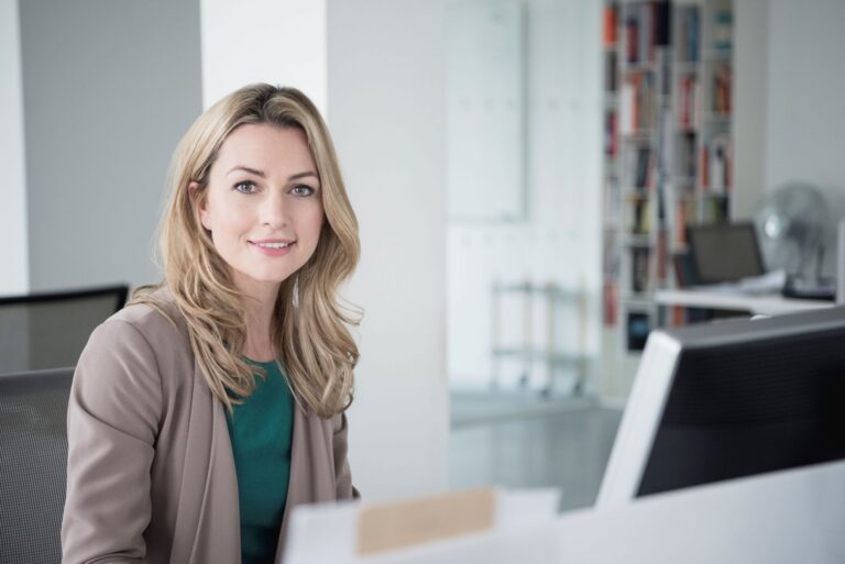 Eine lächelnde junge Geschäftsfrau mit langen blonden Haaren und einem beigen Blazer sitzt an ihrem Schreibtisch in einem hellen, modernen Büro und blickt direkt in die Kamera.