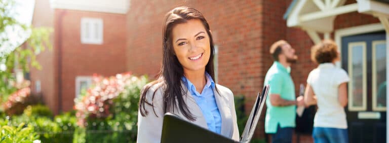 Eine lächelnde junge Immobilienmaklerin steht im Vordergrund, hält eine schwarze Mappe und blickt in die Kamera. Im unscharfen Hintergrund besichtigt ein Paar ein Einfamilienhaus mit roter Ziegelfassade, was durch helles Sonnenlicht hervorgehoben wird.