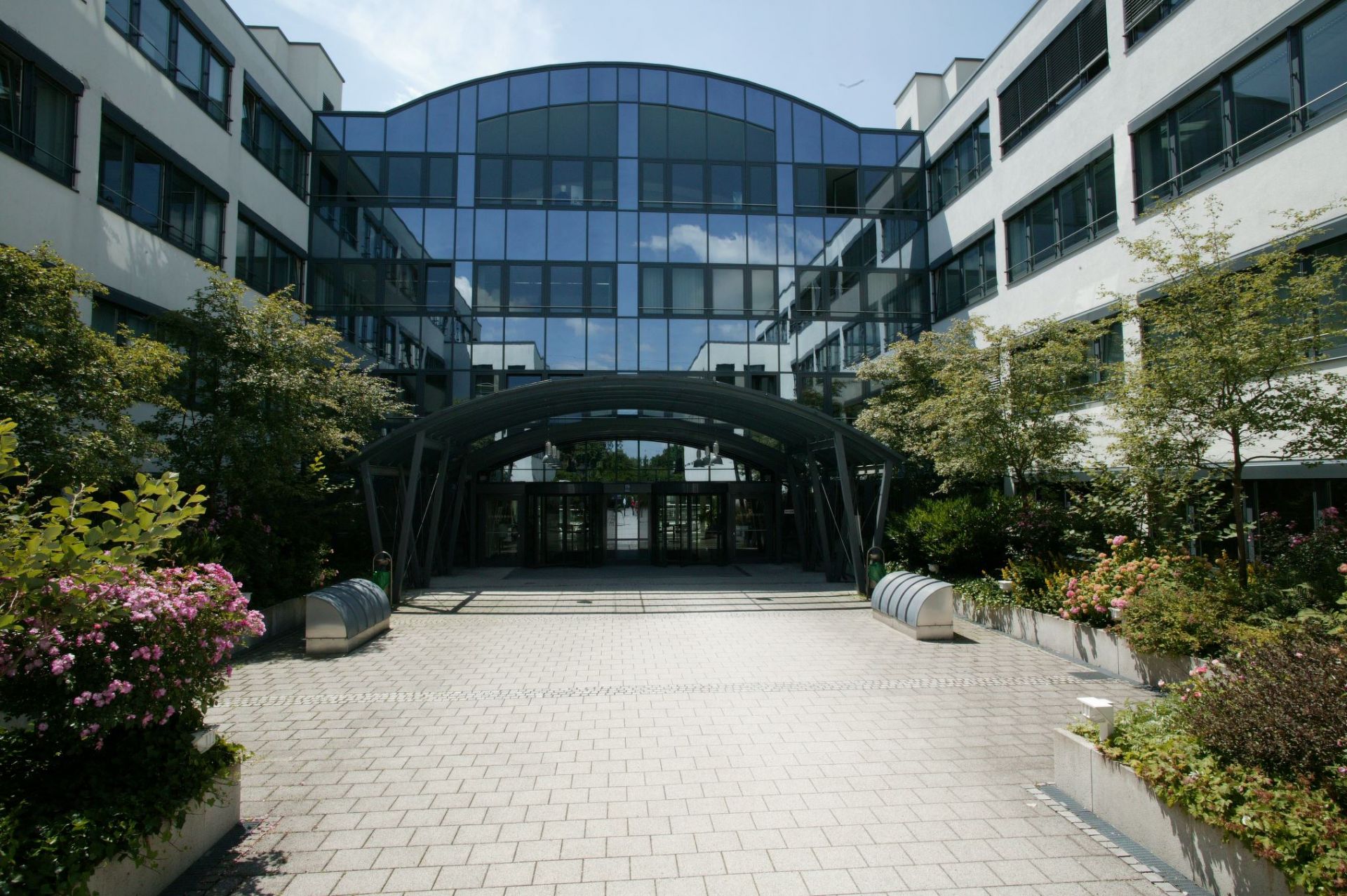 Die moderne Fassade eines Bürogebäudes mit einem halbrunden, verglasten Eingangsbereich und einem davor liegenden gepflasterten Vorplatz. Der Eingang ist von üppigem Grün und Blumenbeeten eingerahmt.