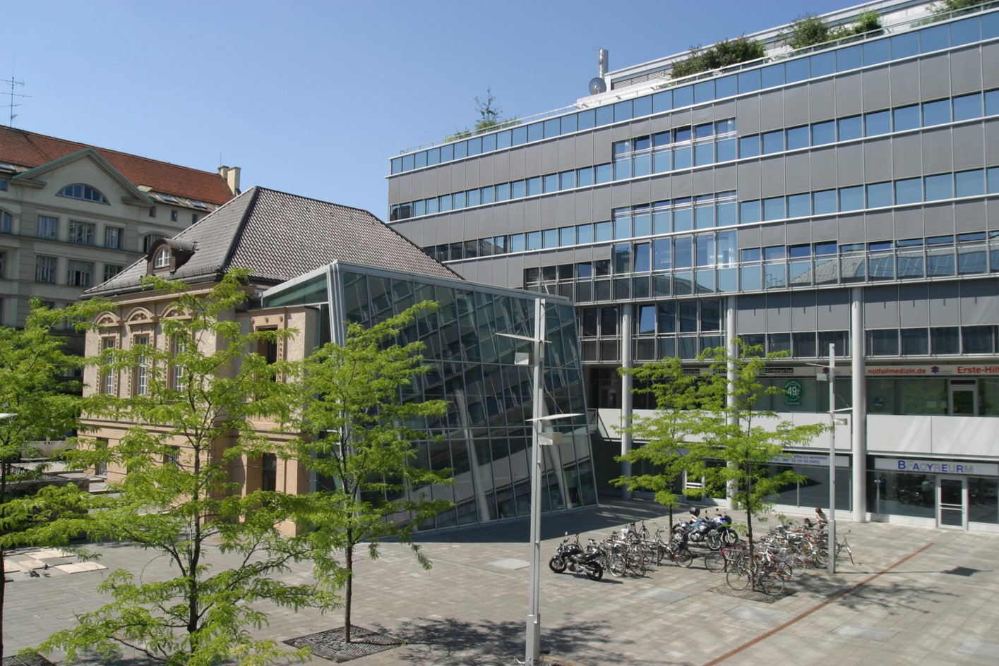 Kontrast zwischen Architektur: Ein modernes, großes Bürogebäude mit grauer Fassade und Glasfront steht rechts neben einem kleineren, älteren, hellbraunen Haus mit traditionellem Dach. Dazwischen befindet sich ein gläserner Verbindungsbau. Im Vordergrund liegt ein gepflasterter Platz mit kleinen Bäumen und Fahrradständern.