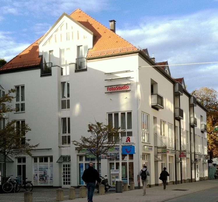 Drei- bis vierstöckiges Eckgebäude in einer Stadtstraße mit weißer Fassade und einem orangefarbenen Ziegeldach. Im Erdgeschoss befinden sich mehrere Geschäfte, darunter eine Apotheke und ein Fotostudio.