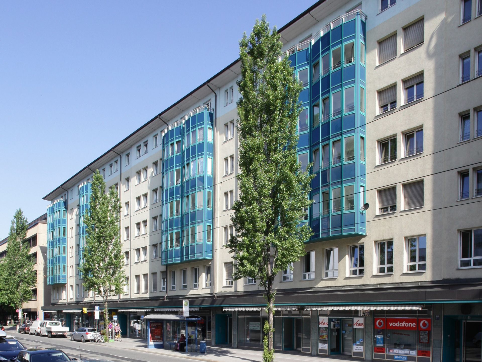 Langes, mehrstöckiges Wohn- und Geschäftshaus mit einer hellen Fassade. Die Gebäudemitte weist vertikale Abschnitte mit hellblauen, verglasten Balkonen auf, die sich von den weißen Fensterreihen abheben. Im Erdgeschoss befinden sich kleine Geschäfte, darunter ein Vodafone-Laden.