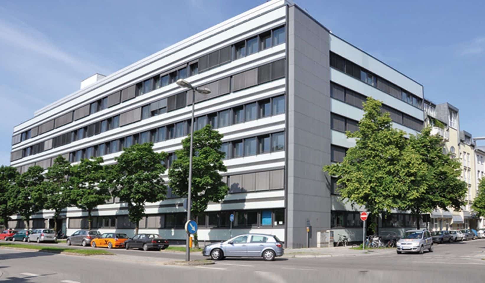 Großes, fünfstöckiges Büro- oder Verwaltungsgebäude mit moderner grauer und weißer Fassade und durchgehenden horizontalen Fensterreihen. Das Gebäude liegt an einer belebten Straßenecke mit parkenden Autos und Bäumen am Straßenrand.