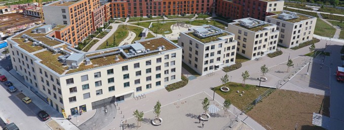 Luftaufnahme einer modernen Wohnsiedlung, bestehend aus einem großen, U-förmigen, rotbraunen Gebäudekomplex und vier kleineren, würfelförmigen, weißen Gebäuden. Alle Flachdächer sind begrünt. In der Mitte befindet sich ein gestalteter Innenhof mit Wegen und Grünflächen.