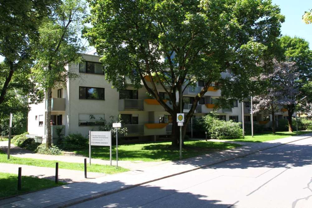Drei- bis vierstöckiges Wohnhaus mit heller Fassade und orangefarbenen Akzenten an den Balkonen, eingebettet in eine grüne Umgebung mit großen Bäumen. Vor dem Gebäude befindet sich ein Schild mit der Aufschrift 'Privates Schülerwohnheim' und 'Leonhard Eißele'.
