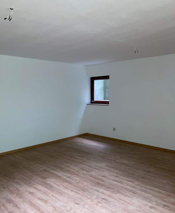 Leerer, neu renovierter Raum mit weißen Wänden, hellem Holzlaminatboden und einem kleinen, dunklen Fenster in der Mitte der Wand.