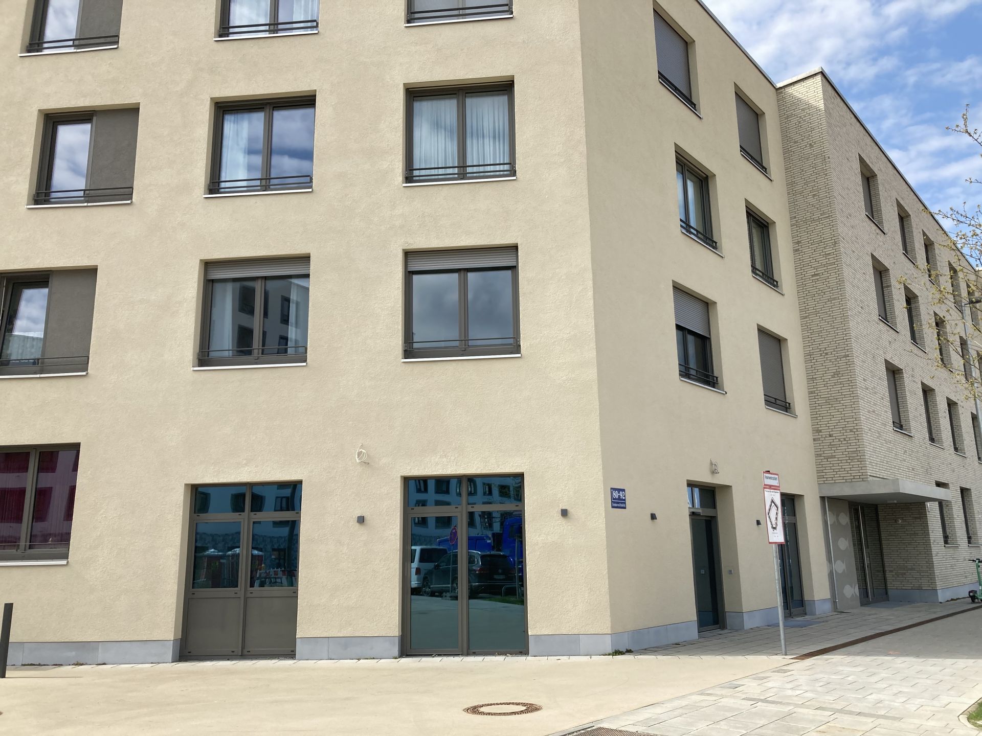Detaillierte Außenansicht der Ecke eines modernen, vierstöckigen Wohn- oder Bürogebäudes mit hellbeiger Putzfassade. Im Erdgeschoss befinden sich zwei gläserne Eingänge, und die Fenster der oberen Stockwerke haben graue Rahmen.