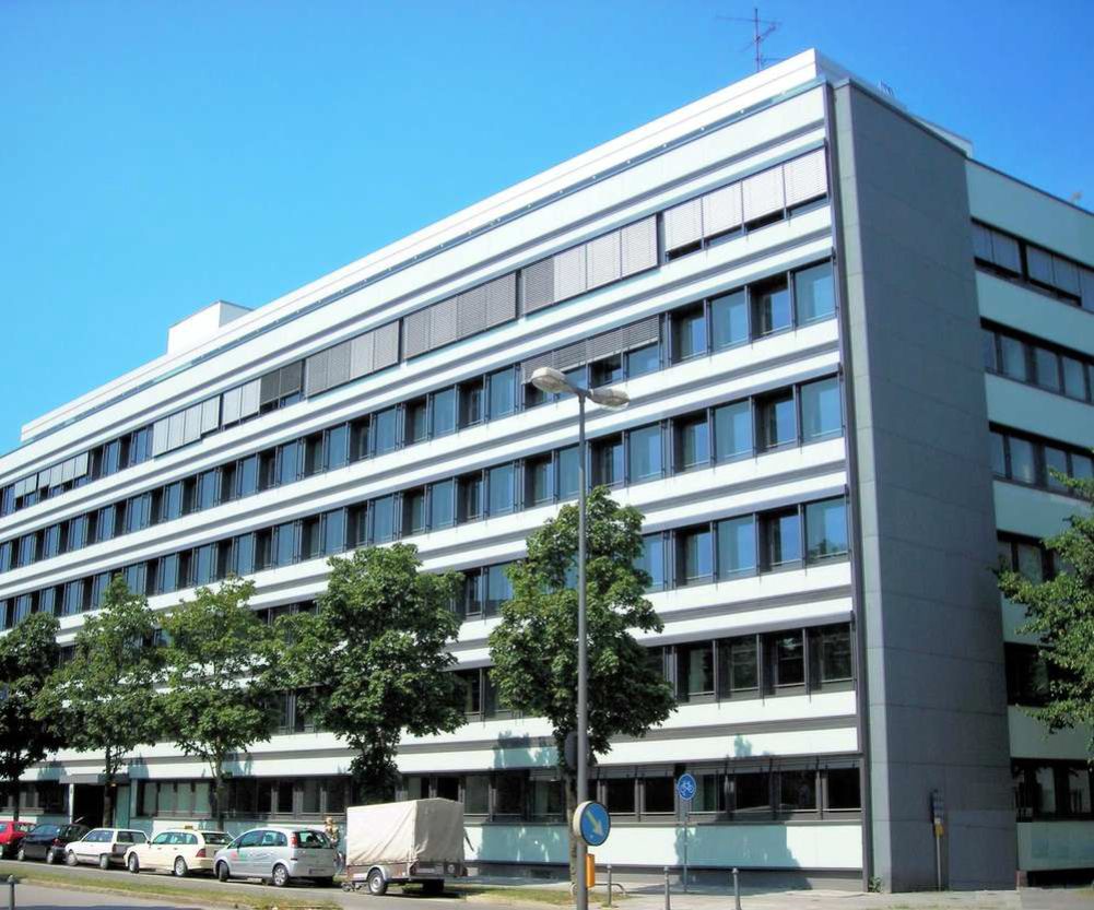 Großes, mehrstöckiges Bürogebäude mit einer Fassade, die abwechselnd aus hellgrauen Streifen und durchgehenden horizontalen Reihen von dunklen Fenstern besteht. Die schmale Seitenfassade ist komplett dunkelgrau. Im Vordergrund stehen eine Reihe von Bäumen, parkende Fahrzeuge und eine Straßenlaterne.