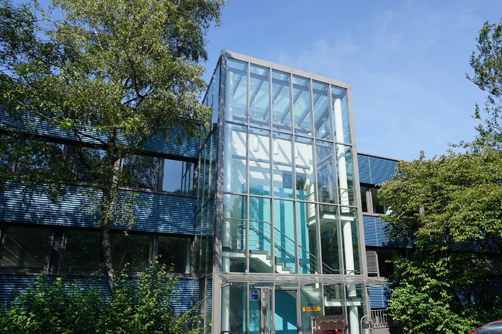 Moderner Eingangsbereich eines Bürogebäudes. Der zentrale, turmartige Eingang besteht aus einer hohen Glasstruktur, die eine interne Treppe und hellblaue Wände umschließt. Die restliche Fassade des Gebäudes ist blau gestreift und von üppigem Grün umgeben.