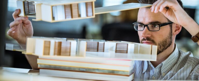 Ein Architekt mit Brille hält und untersucht konzentriert ein detailreiches Holzmodell eines modernen Gebäudes, das er gerade zusammenbaut oder zerlegt.
