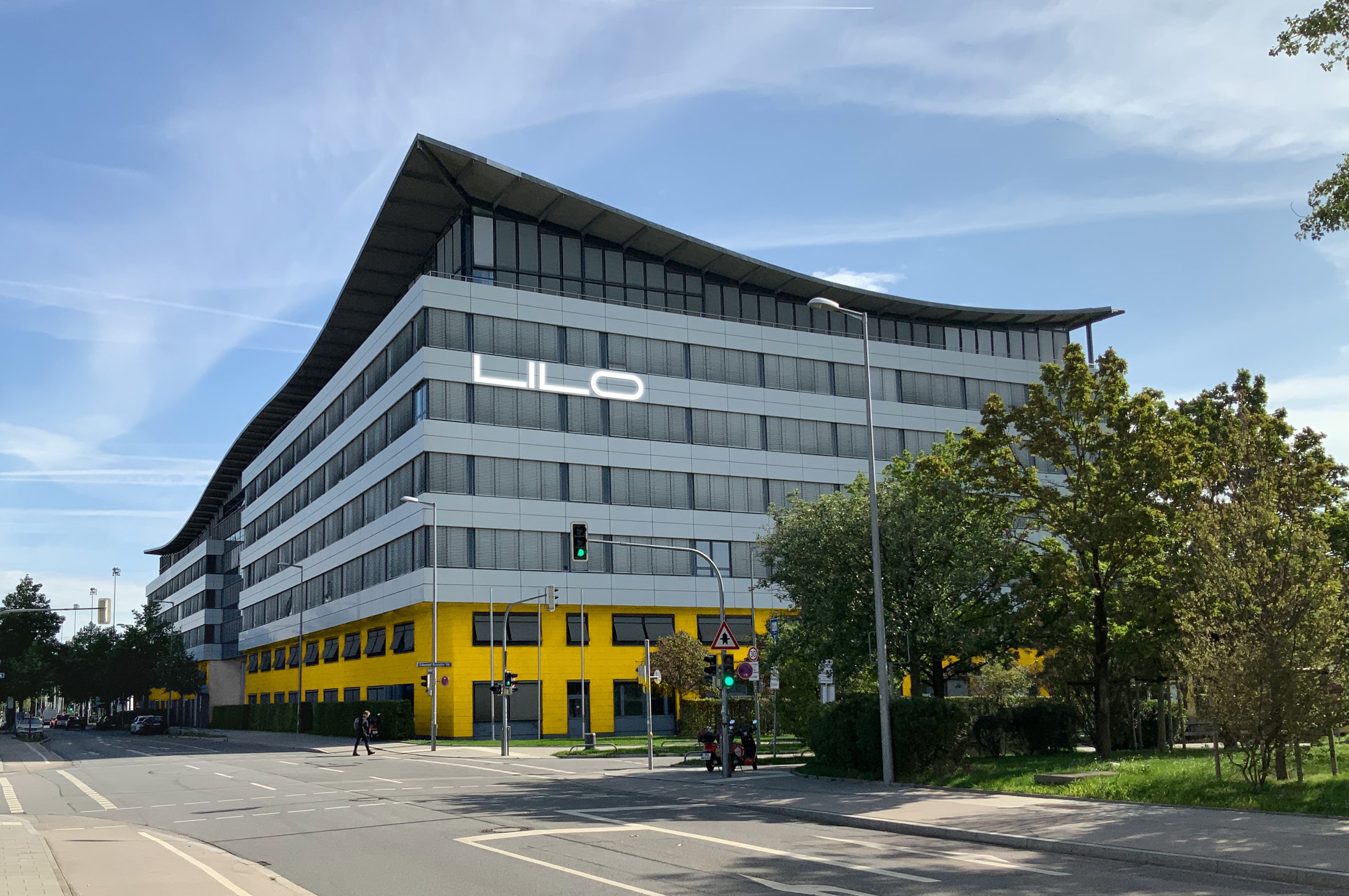 Modernes, fünfstöckiges Bürogebäude an einer Straßenecke mit einer geschwungenen Dachlinie. Die unteren beiden Etagen sind leuchtend gelb gestrichen, die oberen drei Etagen haben eine graue Glasfassade. Der Schriftzug 'LILO' ist in Großbuchstaben auf der Fassade sichtbar.